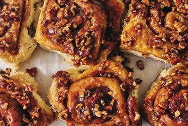 Overhead view of a tray of dark brown sticky buns fresh out of the oven and covered in nuts and glaze.