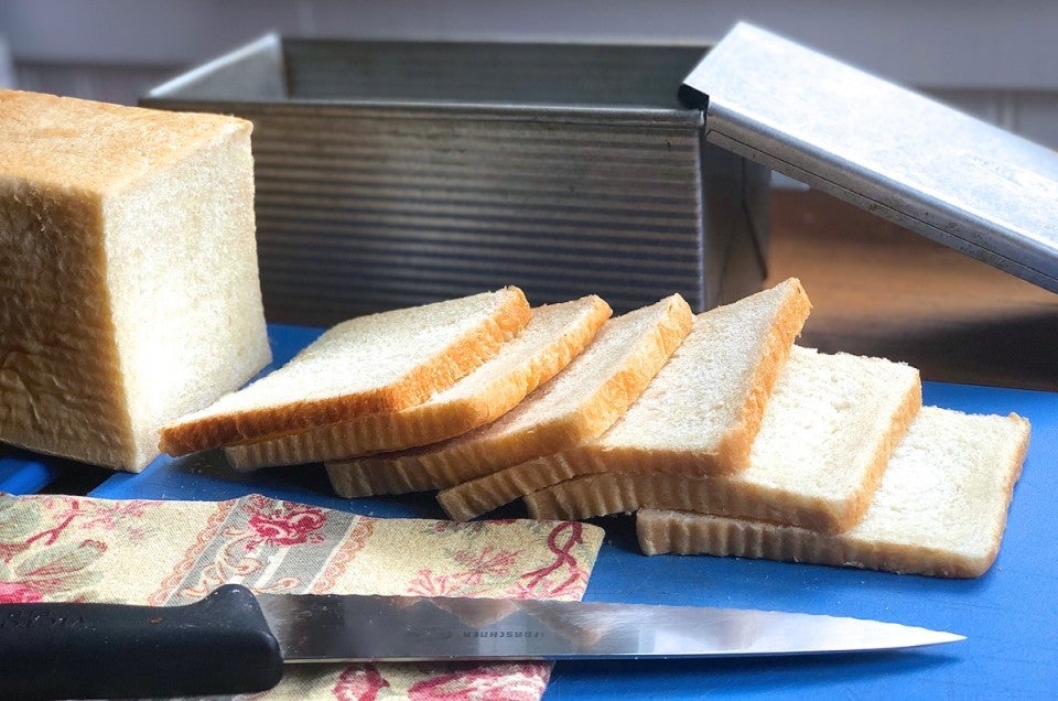 Loaf of white sandwich bread, sliced, on a cutting board in front of a pain de mie pan.