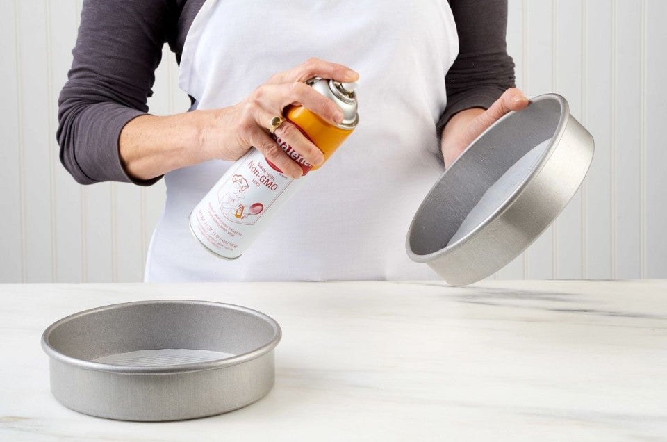 Round cake pans, lined with parchment, being sprayed with nonstick vegetable oil spray.