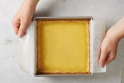 Hands using parchment paper sling to remove lemon bars from square pan