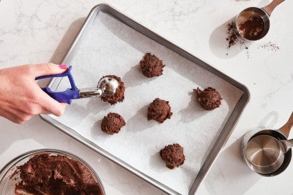 Chocolate cookie dough being scooped and spaced several inches apart on a parchment-lined baking sheet