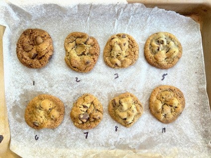 Chocolate chip cookies on a parchment-lined baking sheet, showing their varying degrees of browning based on what air fryer setting they were baked on.