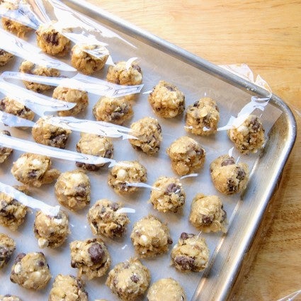 Balls of chocolate chip cookie dough lined up on a baking sheet and covered with plastic wrap, ready to be frozen and then bagged. 