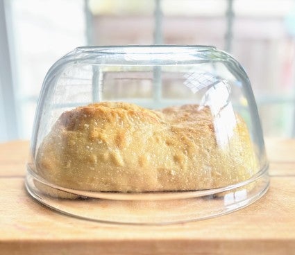 Oval loaf of sourdough bread resting on a cutting board beneath a glass bowl.